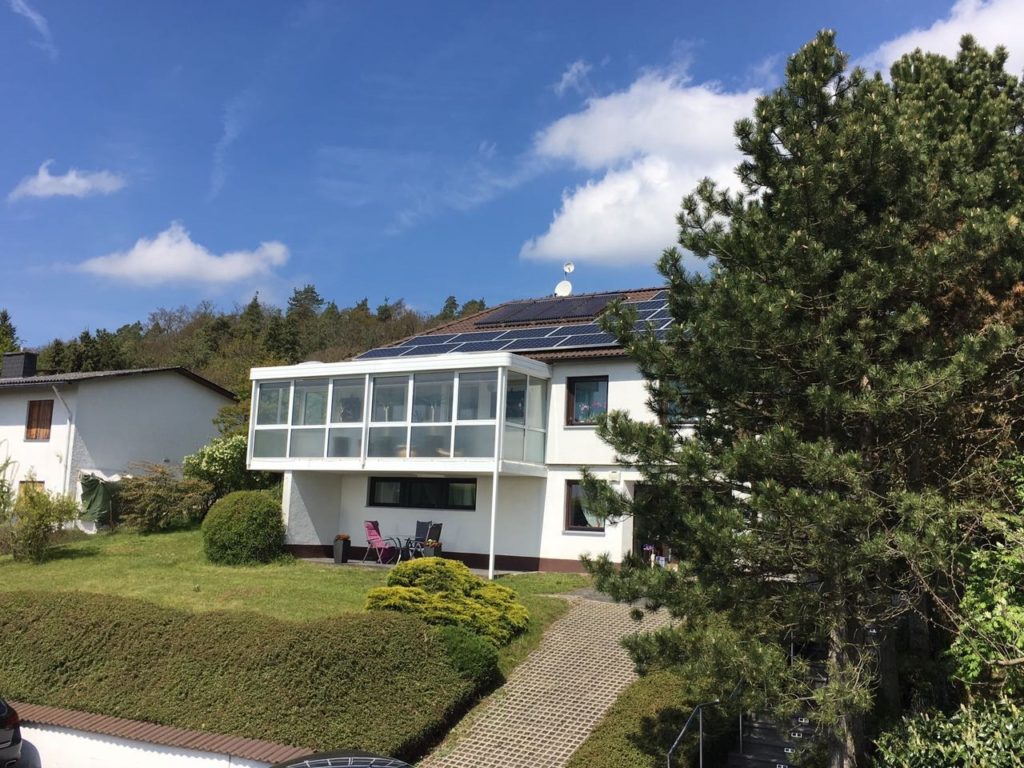 Photovoltaikanlage auf dem Dach eines weißen Einfamilienhauses mit Wintergarten, Terrasse und großem Garten bei sonnigem Wetter
